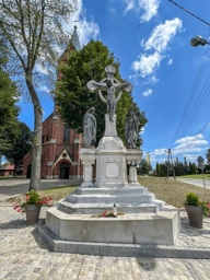 A stone crucifix monument with two figures, surrounded by potted plants, with a brick church and trees in the background.