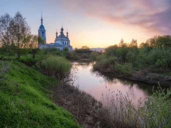 A picturesque view of a church by a river during sunset, surrounded by greenery.